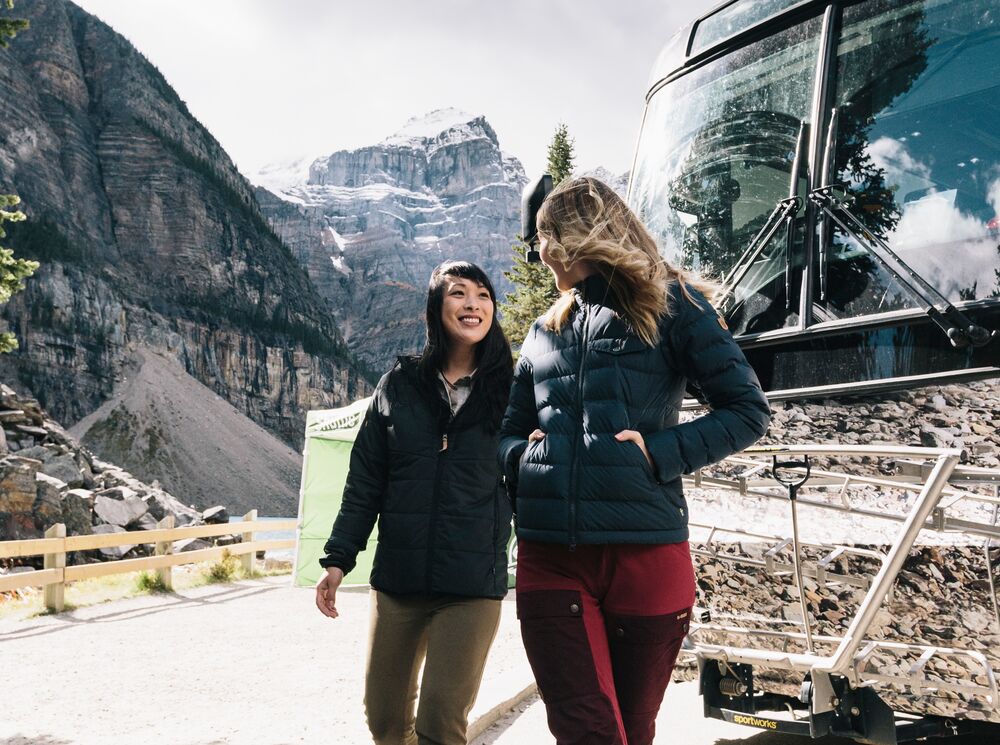 Two people in front of the Roam Public Transit bus at Moraine Lake on a sunny fall day in Banff National Park.